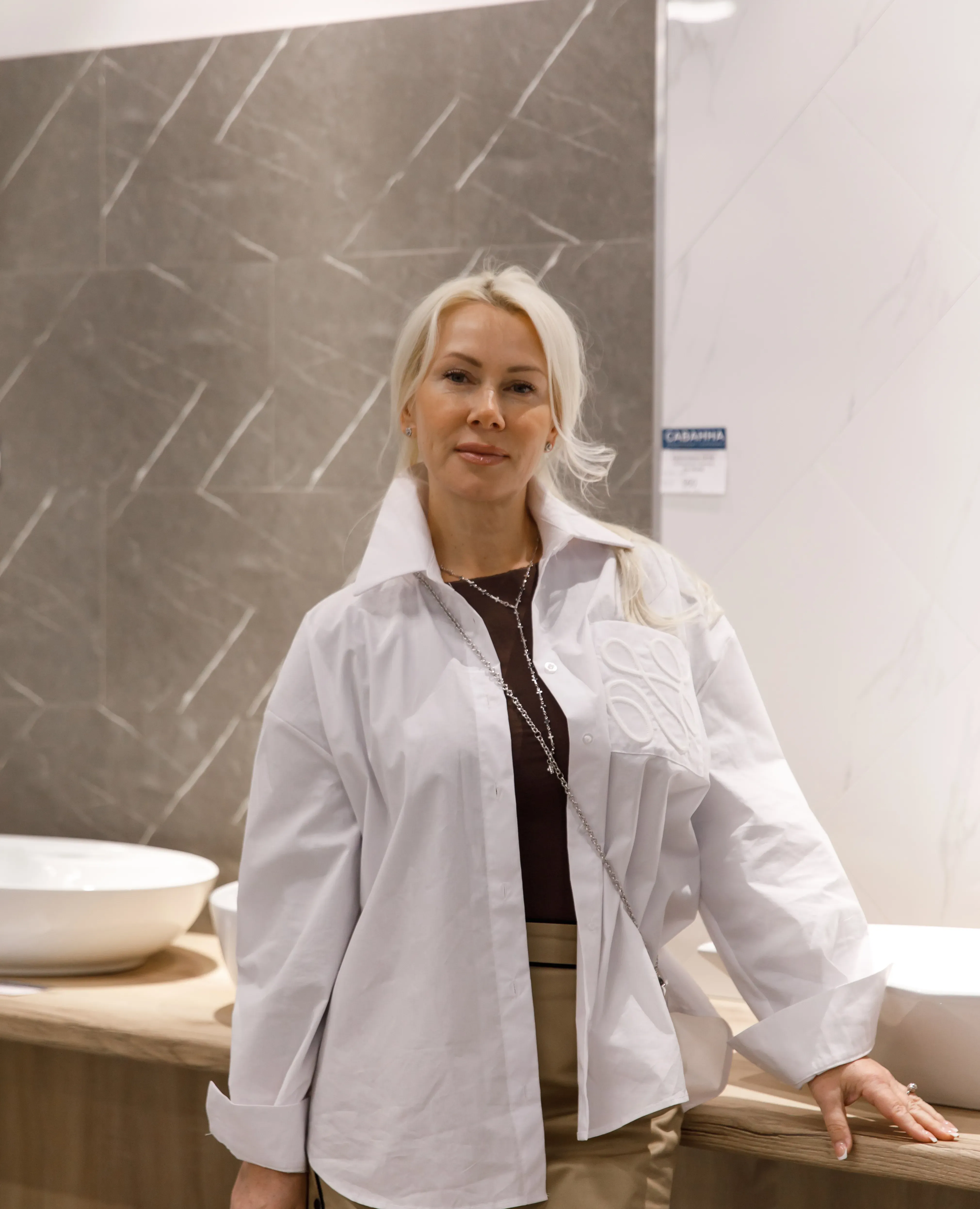 A blonde woman in a white shirt stands in a modern bathroom showroom with sinks.